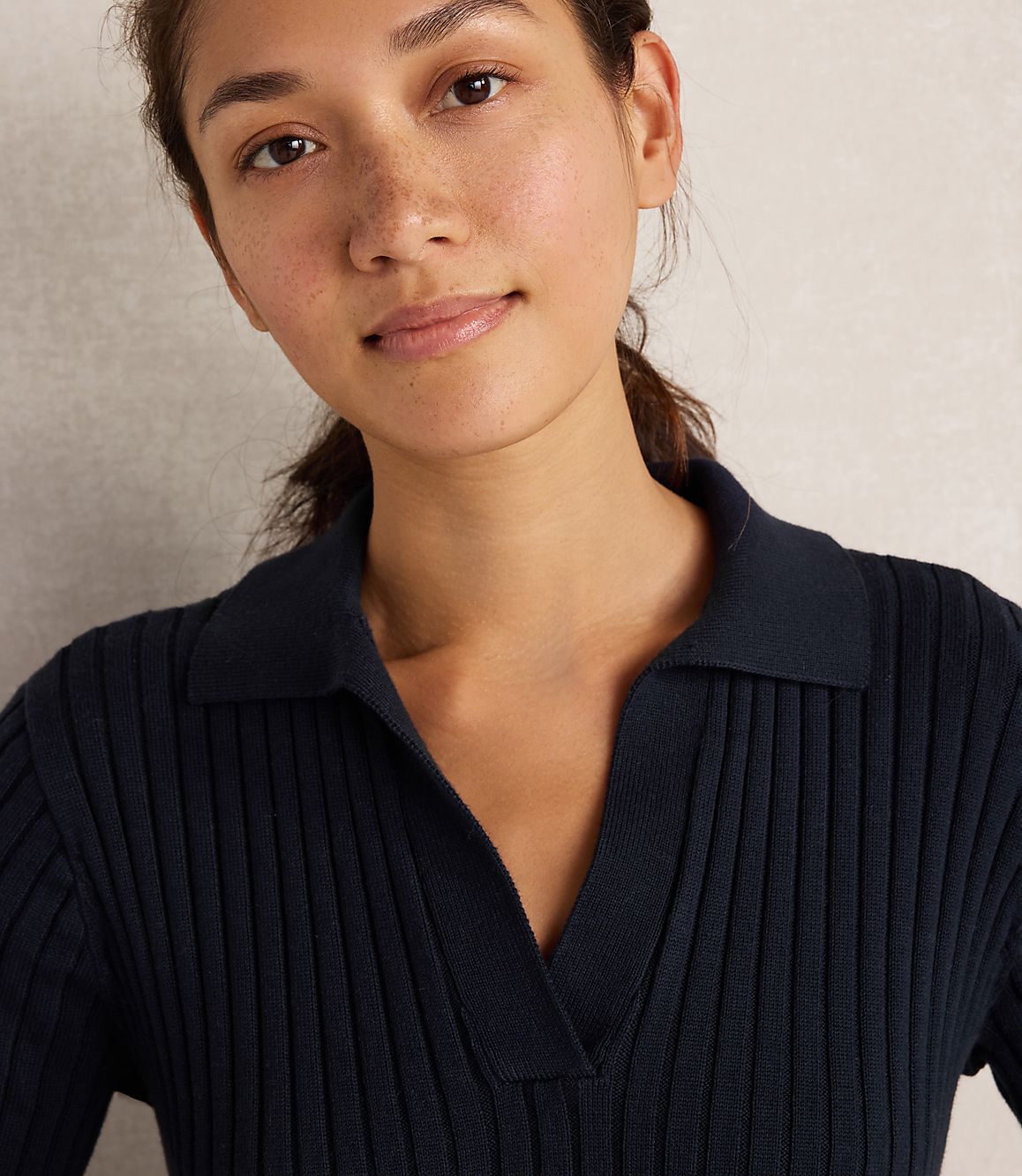A young woman with dark hair pulled back smiles gently in a dark blue ribbed polo shirt against a neutral, textured background.