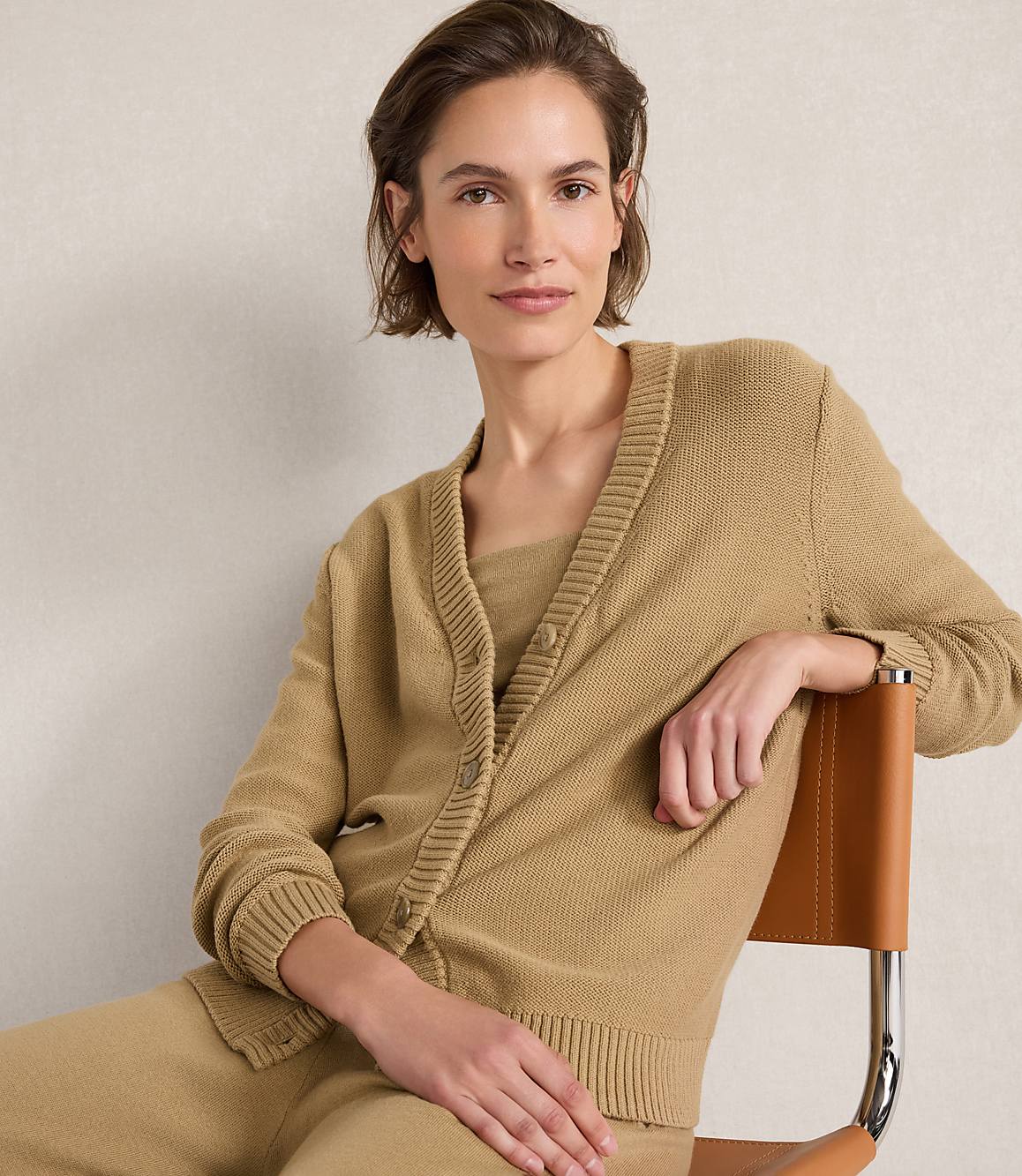 A woman in a tan cardigan and matching top sits on a chair with a light-colored wall behind her.