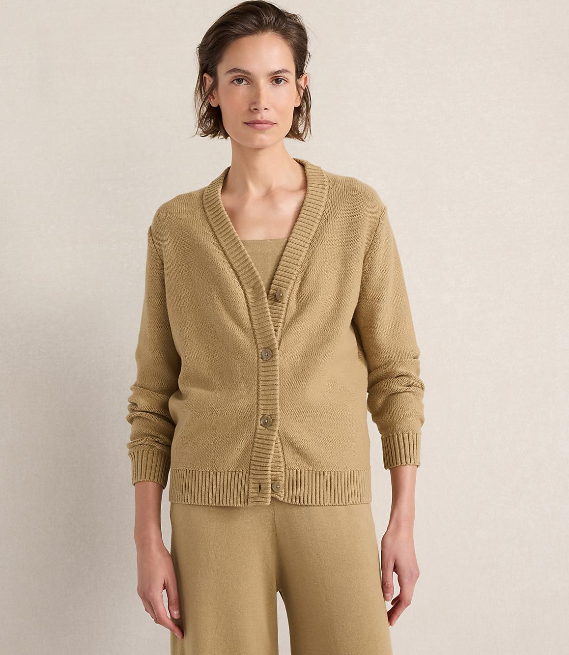 A woman in a tan cardigan and matching pants stands against a plain, light-colored background.