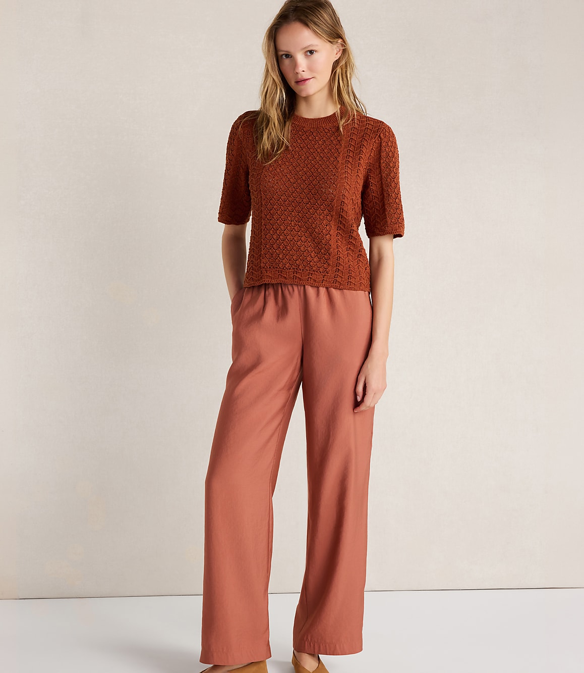 A woman in a rust-colored textured top and wide-leg rust pants stands against a textured cream background.