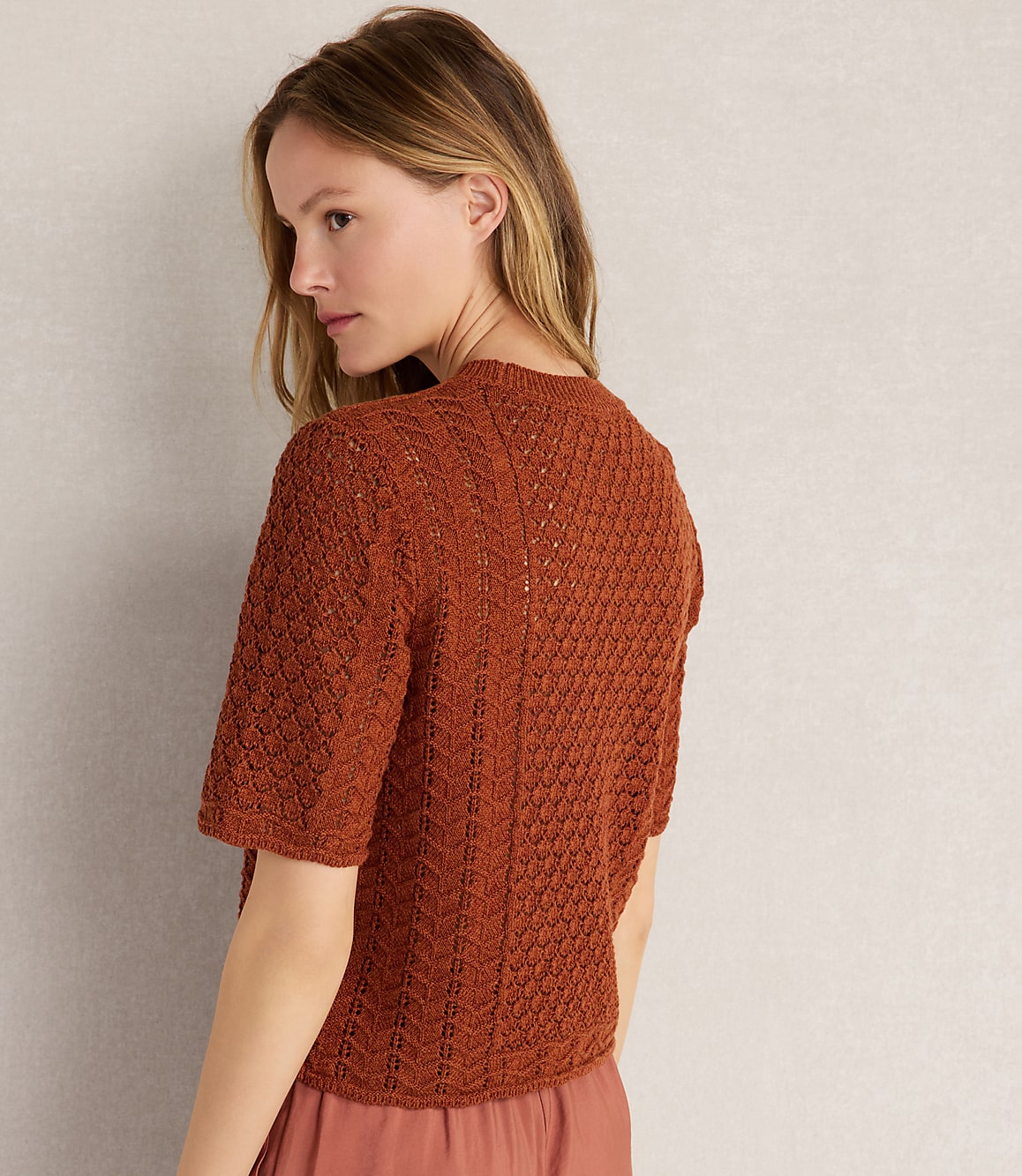 A woman in a textured rust-colored sweater is shown from the back against a plain, light-colored wall.