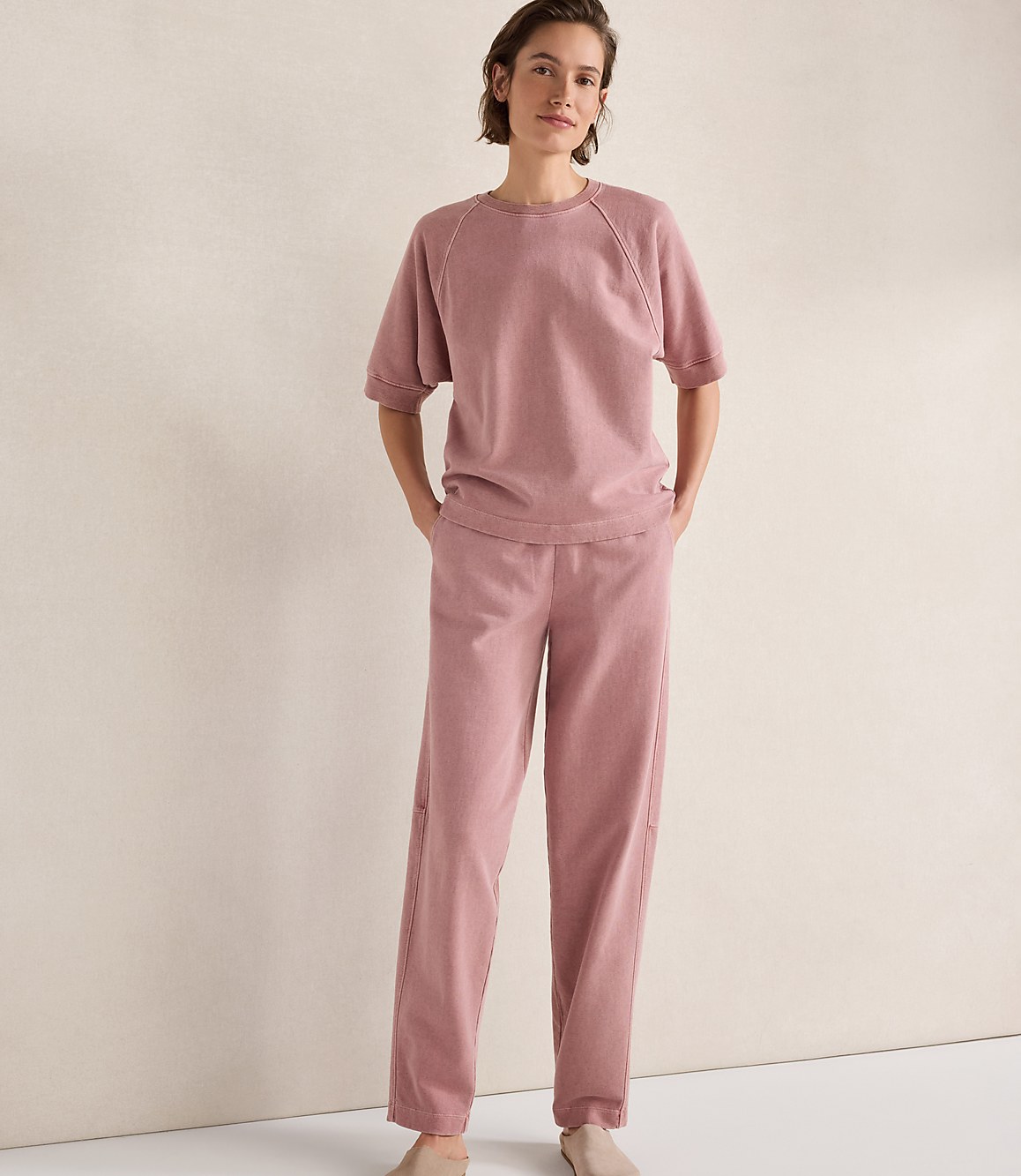 A woman in a pink loungewear set stands with her hands in her pockets against a plain, light-colored wall.