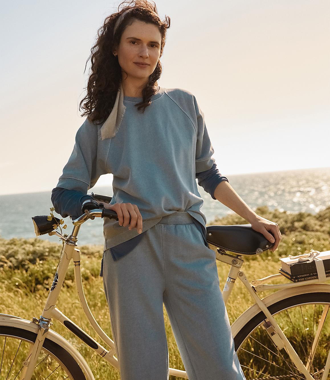 A woman in casual clothing poses with a bicycle in the foreground, while a sunlit ocean and coastline stretch across the background.