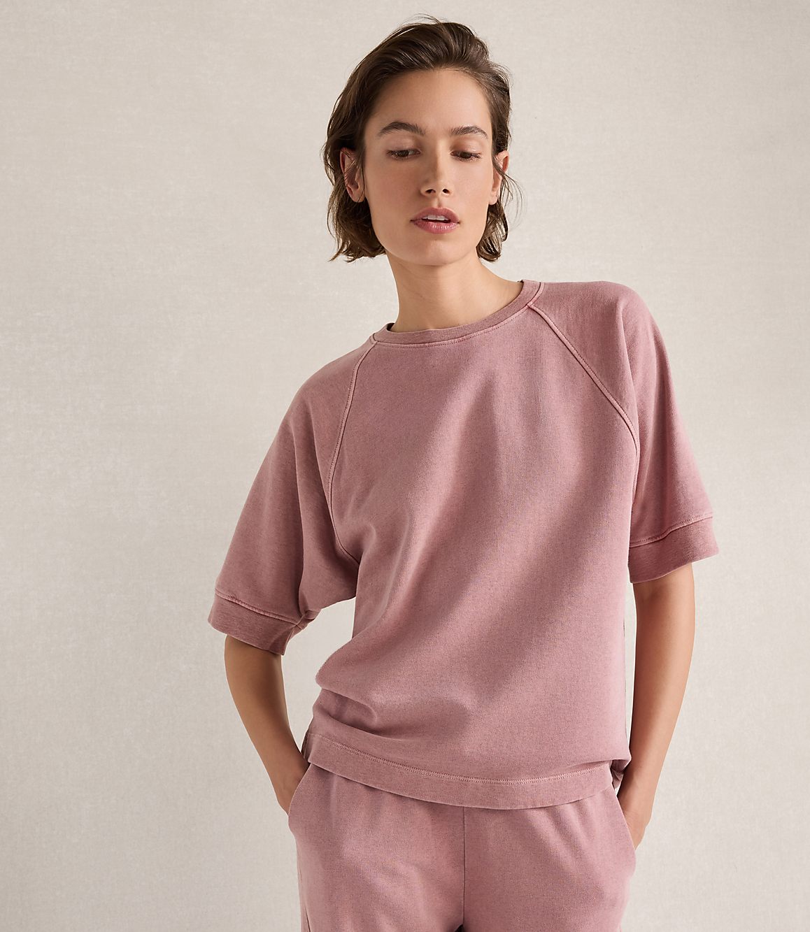 A woman in a pink sweatshirt and matching pants stands against a textured, light-colored wall.