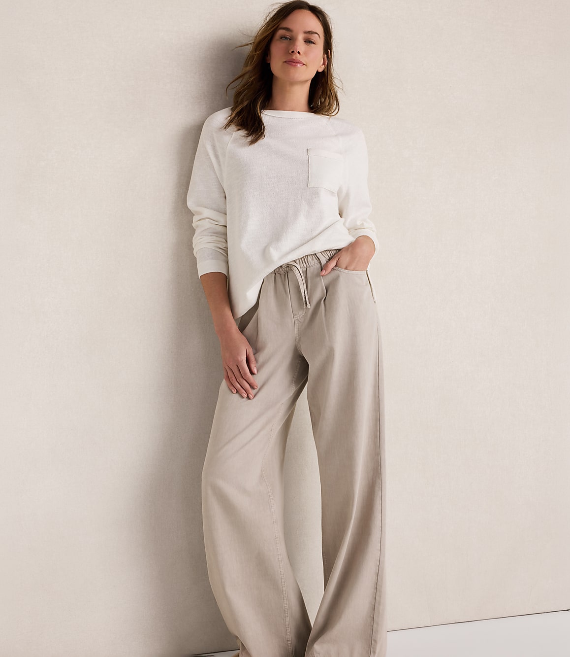 A woman in a white long-sleeved shirt and beige wide-leg pants stands against a textured cream wall.