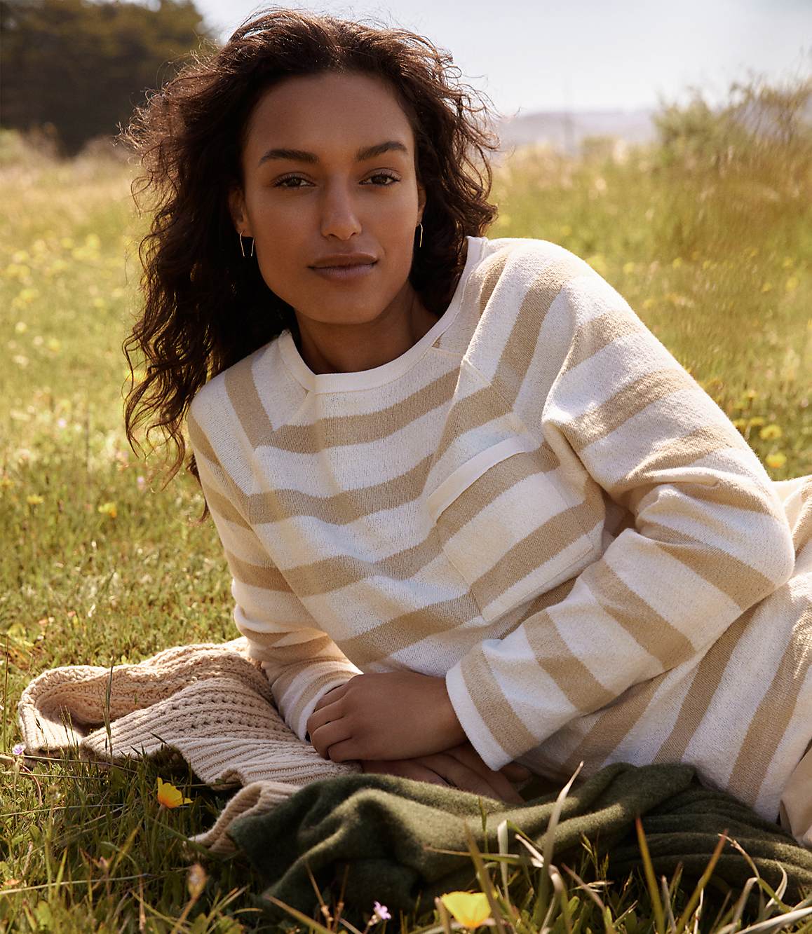 A woman with dark curly hair lies on a blanket in a grassy field, with a blurred background of trees and sky.