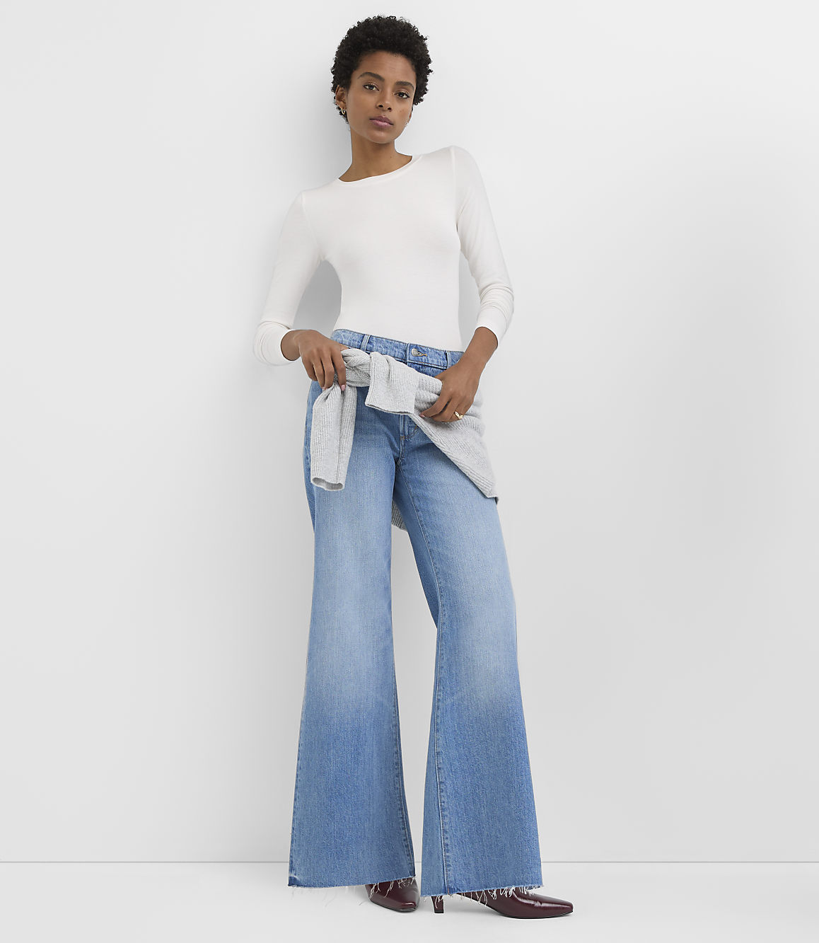 A woman in a white long-sleeved shirt and wide-leg jeans stands against a plain white background.