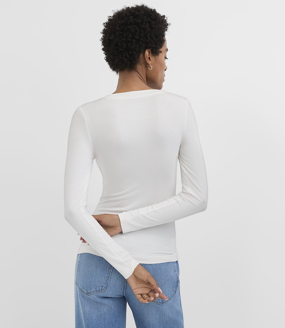 A woman in a white long-sleeved shirt and blue jeans stands with her back to the viewer against a plain white background.