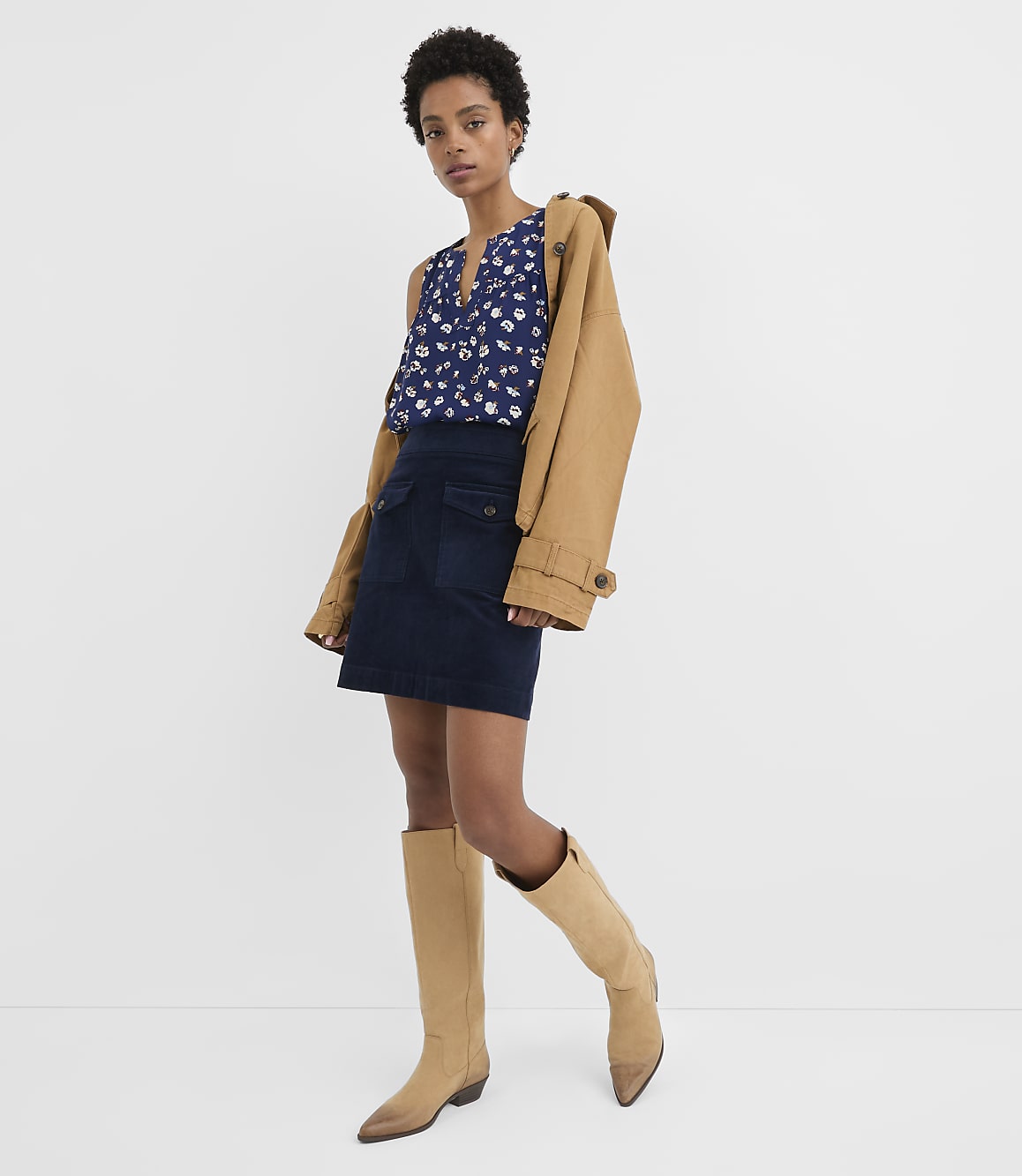 A woman in a floral top, denim skirt, and tan boots stands against a plain white background with a tan jacket draped over her shoulder.