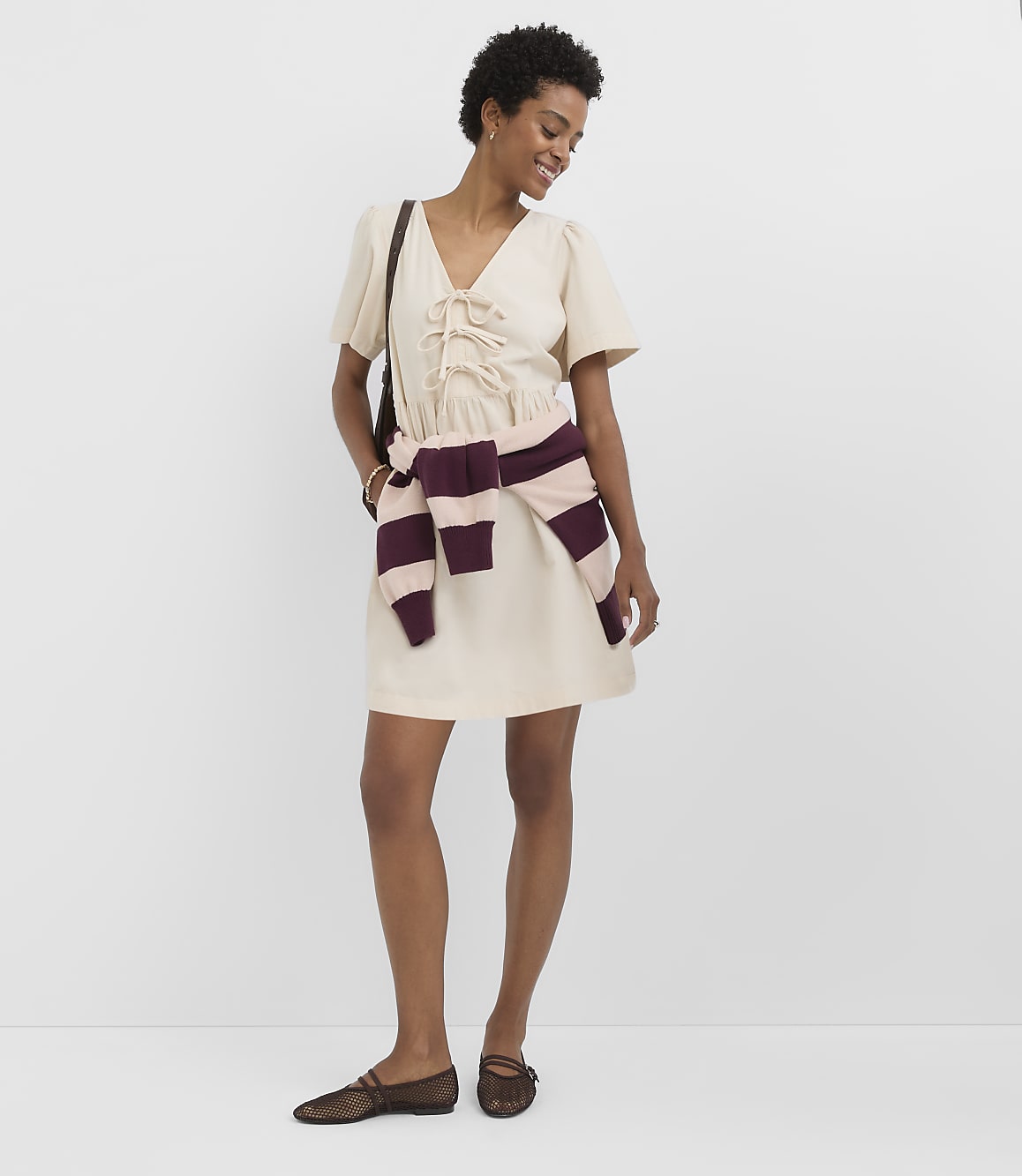 A woman in a cream dress and sheer flats stands against a plain white background, with a striped sweater tied around her waist.