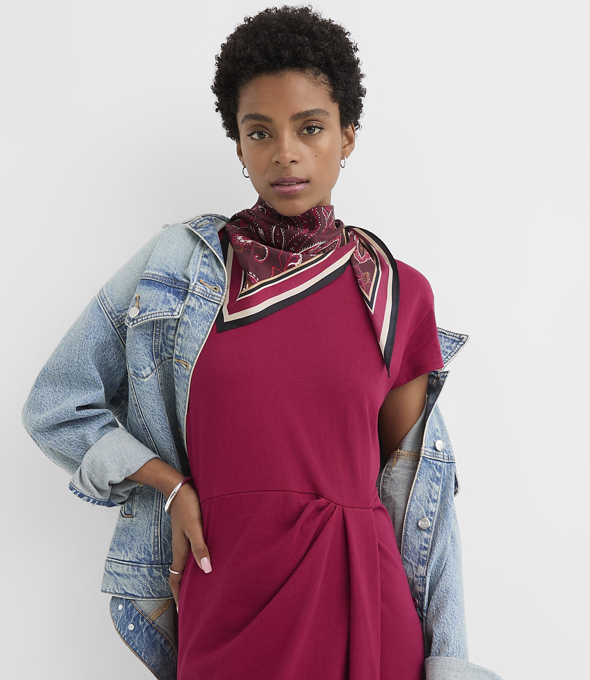 A woman in a maroon dress and denim jacket stands against a plain white background.