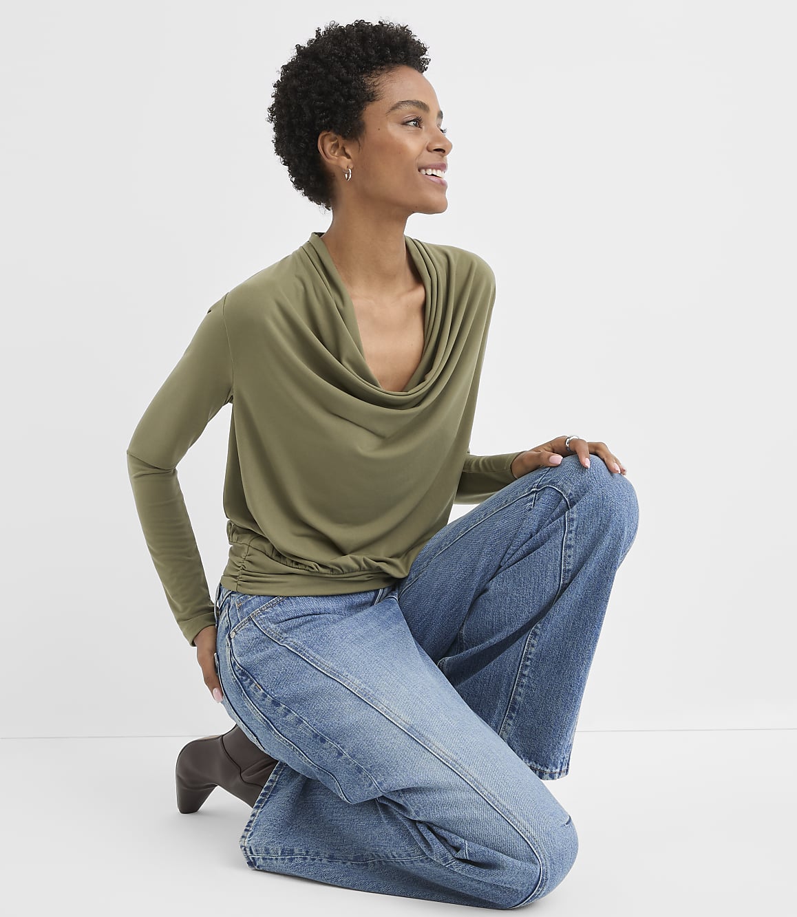 A woman in a green cowl-neck top and blue jeans kneels against a plain white background.