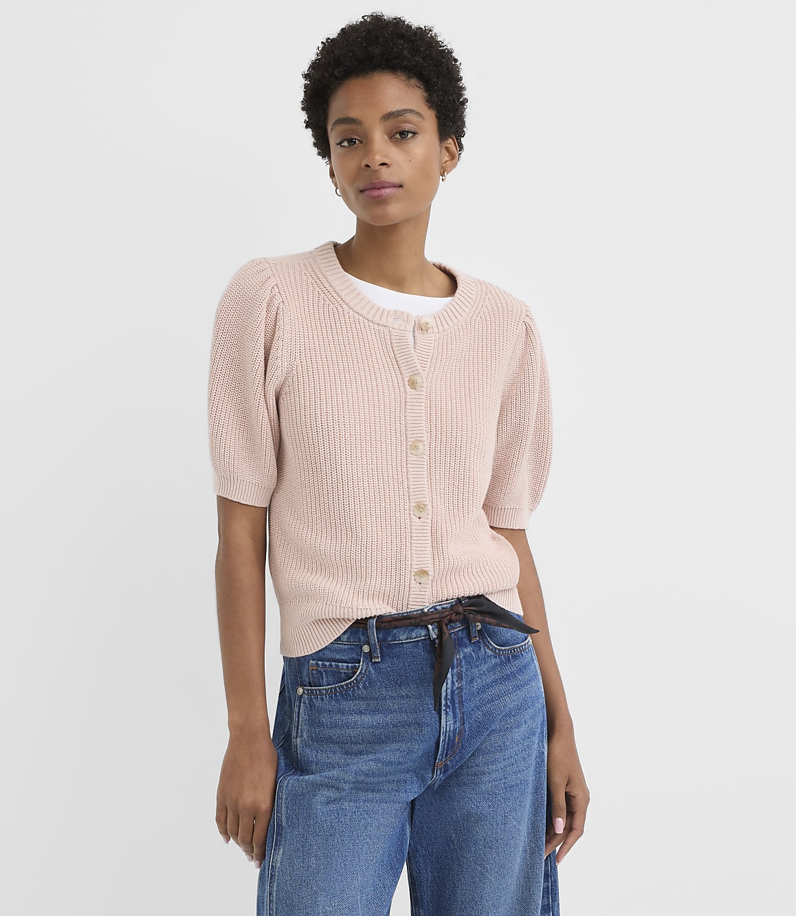A woman in a pink cardigan and blue jeans stands against a plain white background.