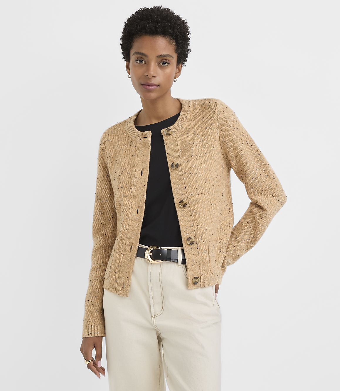 A woman in a tan speckled cardigan and cream pants stands against a plain white background.