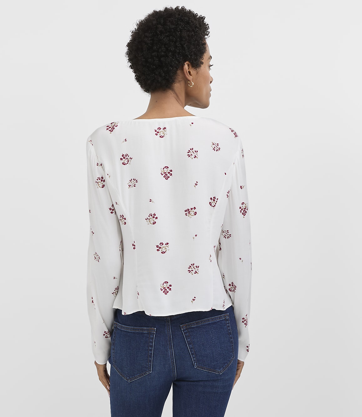 A woman in a white floral blouse and blue jeans stands against a plain white background.