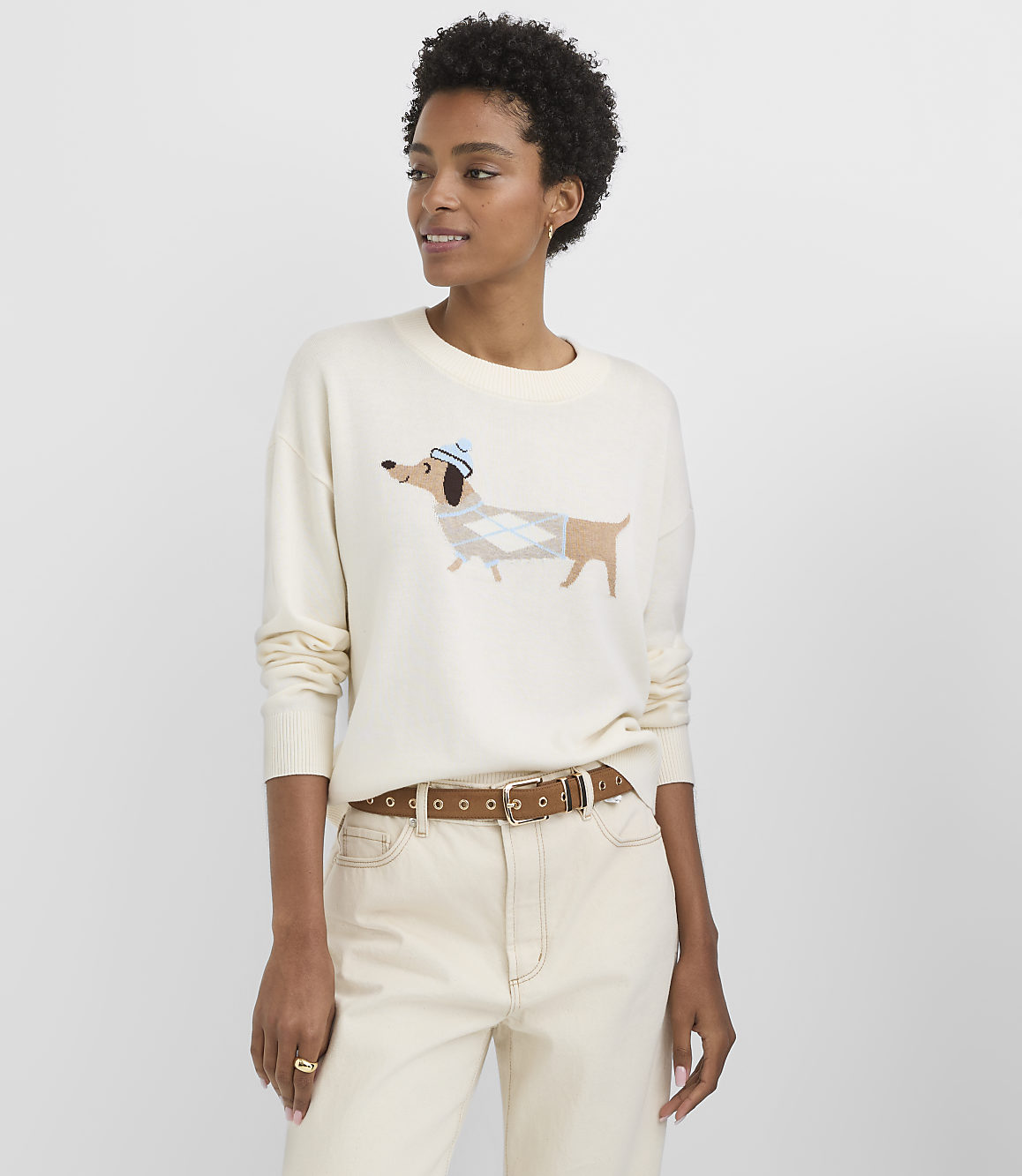 A woman in a cream sweater with a dachshund design and cream pants stands against a plain white background.