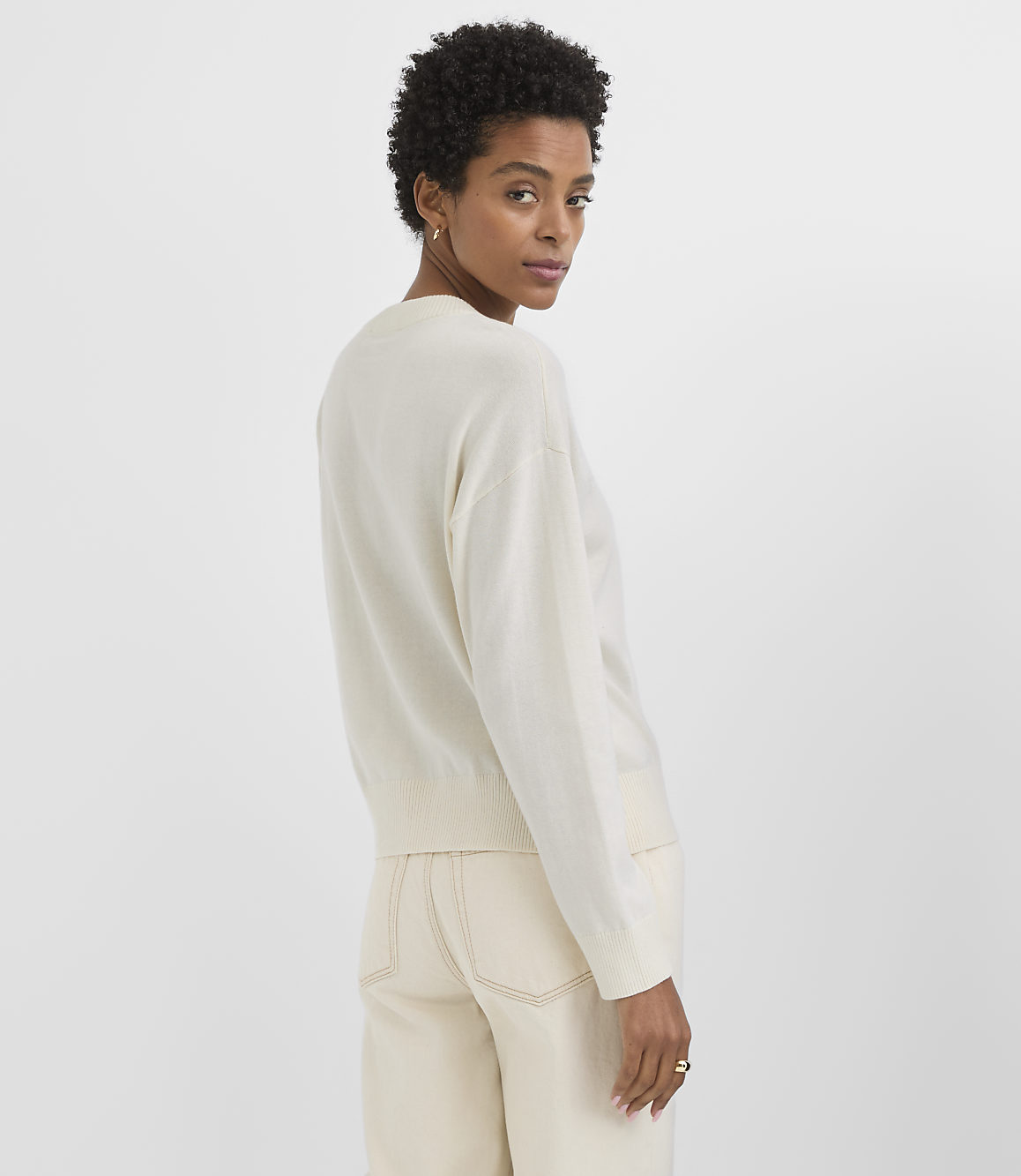 A woman in a cream sweater and pants stands against a plain white background.
