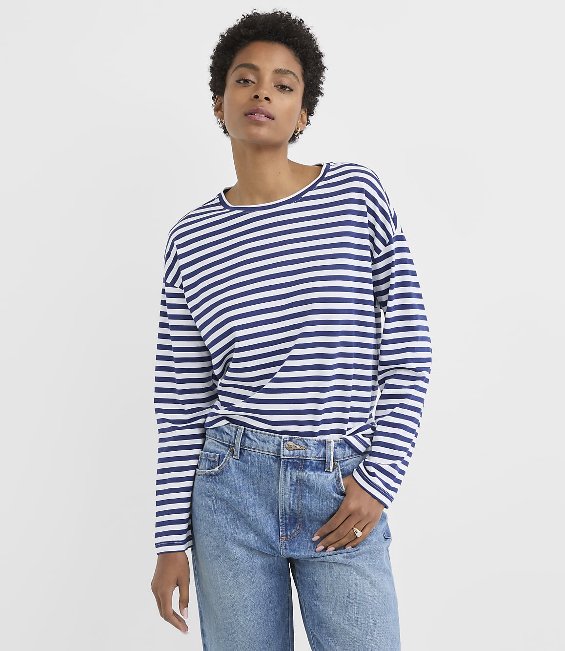 A woman in a striped long-sleeved shirt and jeans stands against a plain white background.