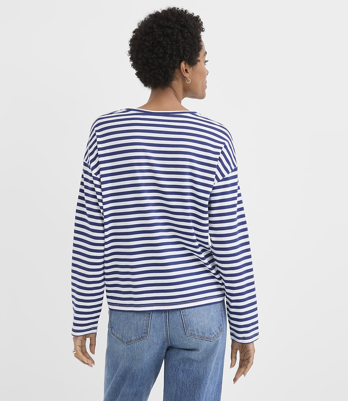 A woman in a striped shirt and jeans stands against a plain white background.