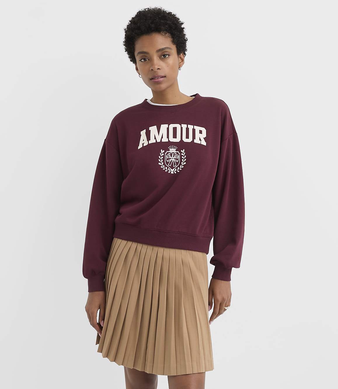 A woman in a maroon sweatshirt with "AMOUR" printed on it and a tan pleated skirt stands against a plain white background.