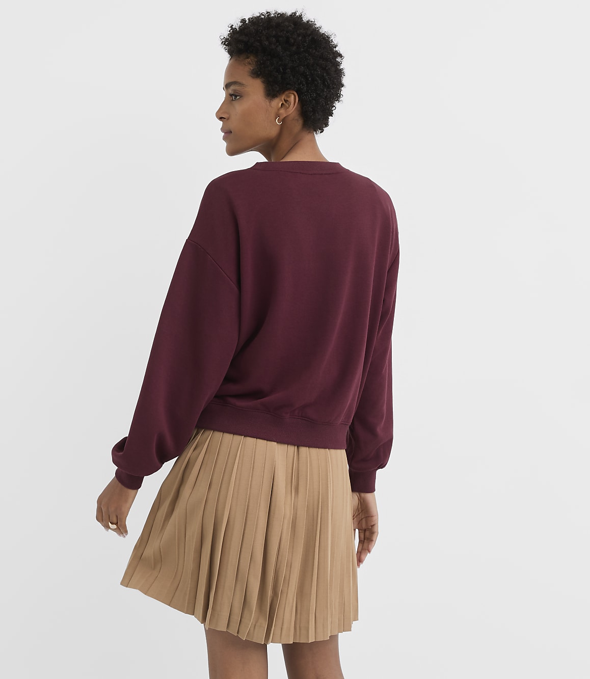 A woman in a maroon sweatshirt and a tan pleated skirt stands against a plain white background.
