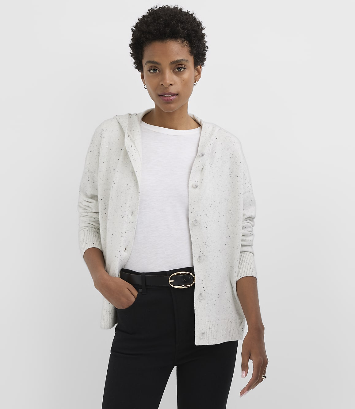 A woman in a light-colored hooded cardigan and black pants stands against a plain white background.