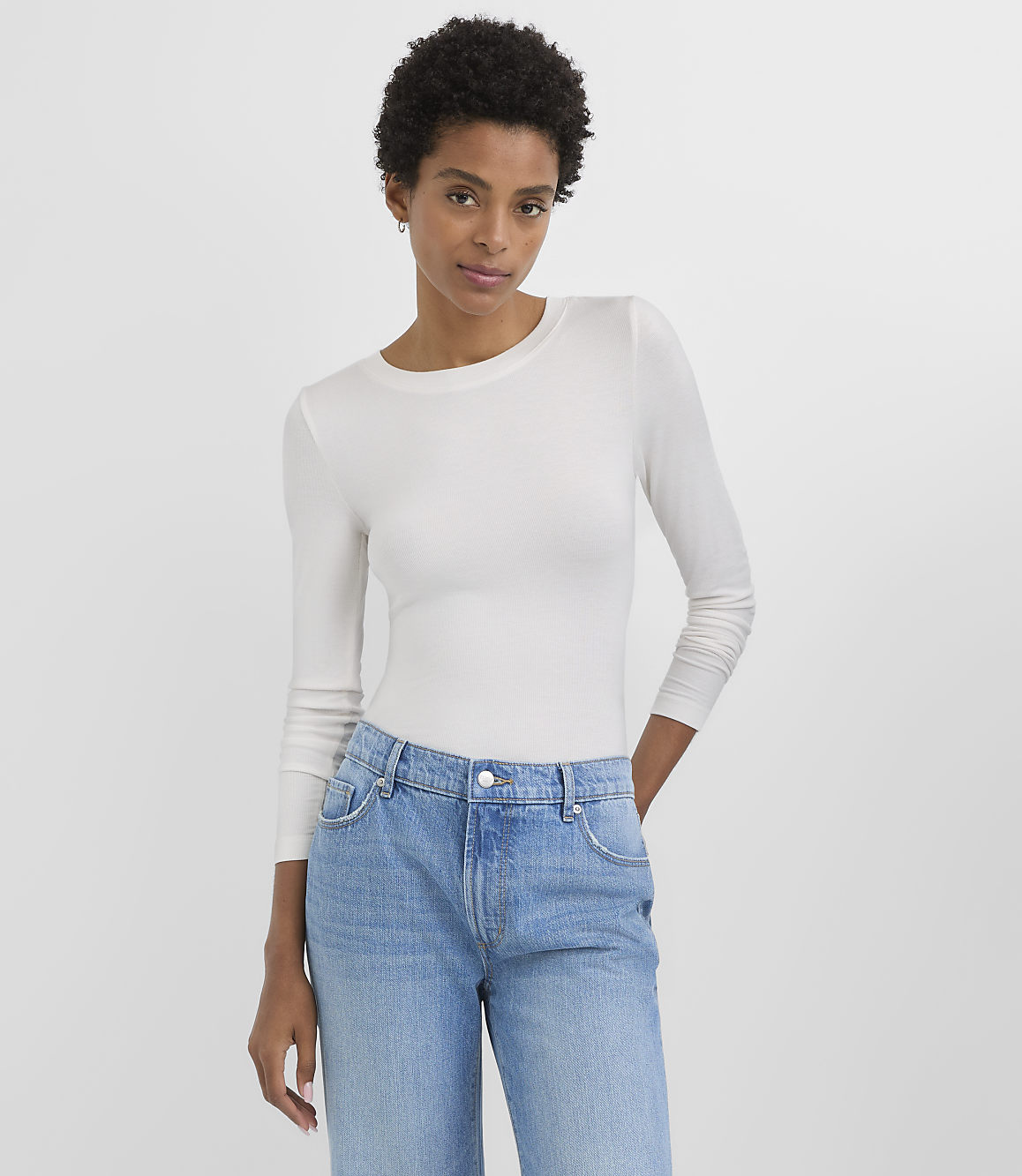 A woman in a white long-sleeved shirt and blue jeans stands against a plain white background.