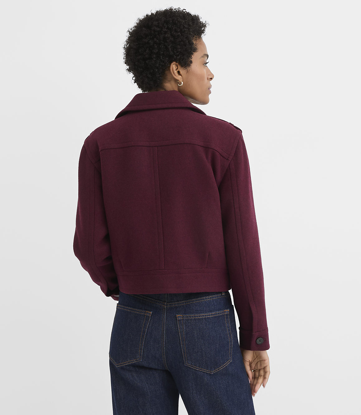 A woman in a maroon jacket and dark jeans stands against a plain white background.