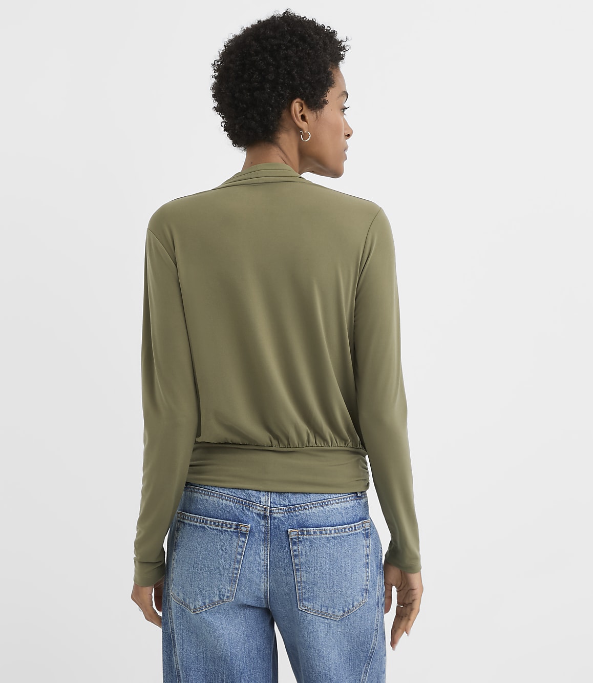 A woman in a green wrap top and blue jeans stands against a plain white background.