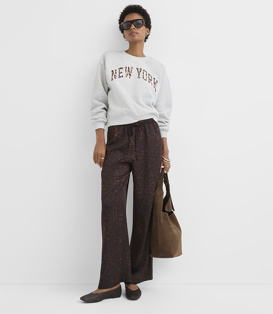A woman in a "New York" sweatshirt and patterned pants stands against a plain white wall, holding a brown bag.