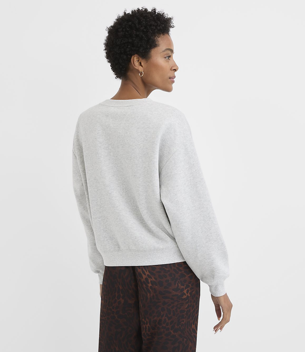A woman in a grey sweatshirt and patterned pants stands against a plain white background.