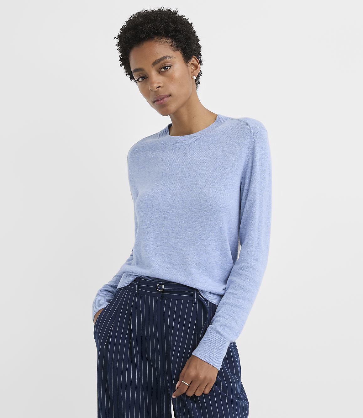 A woman in a light blue sweater and pinstripe pants stands against a plain white background.