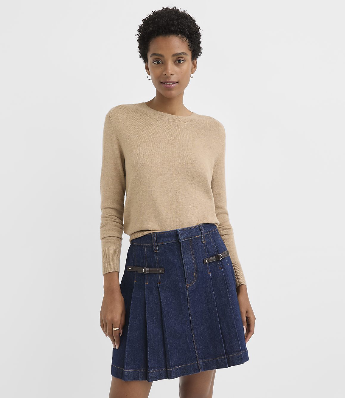 A woman in a tan sweater and denim skirt stands against a plain white background.
