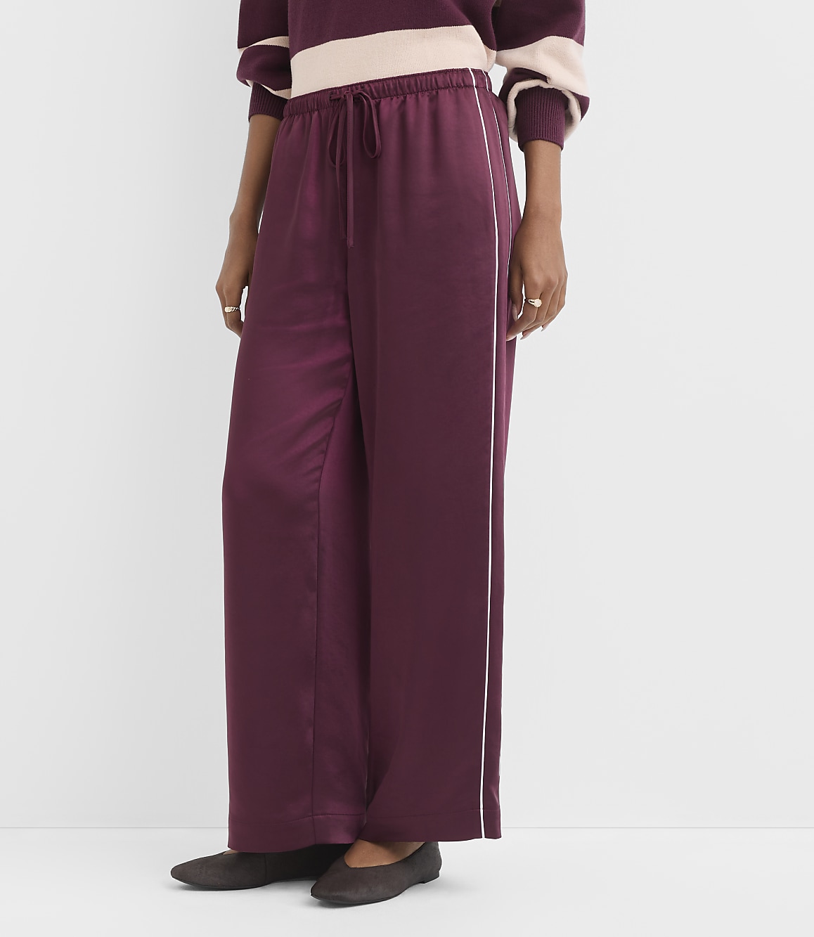 A person wearing burgundy satin wide-leg pants with a white stripe down the side stands against a plain white background.