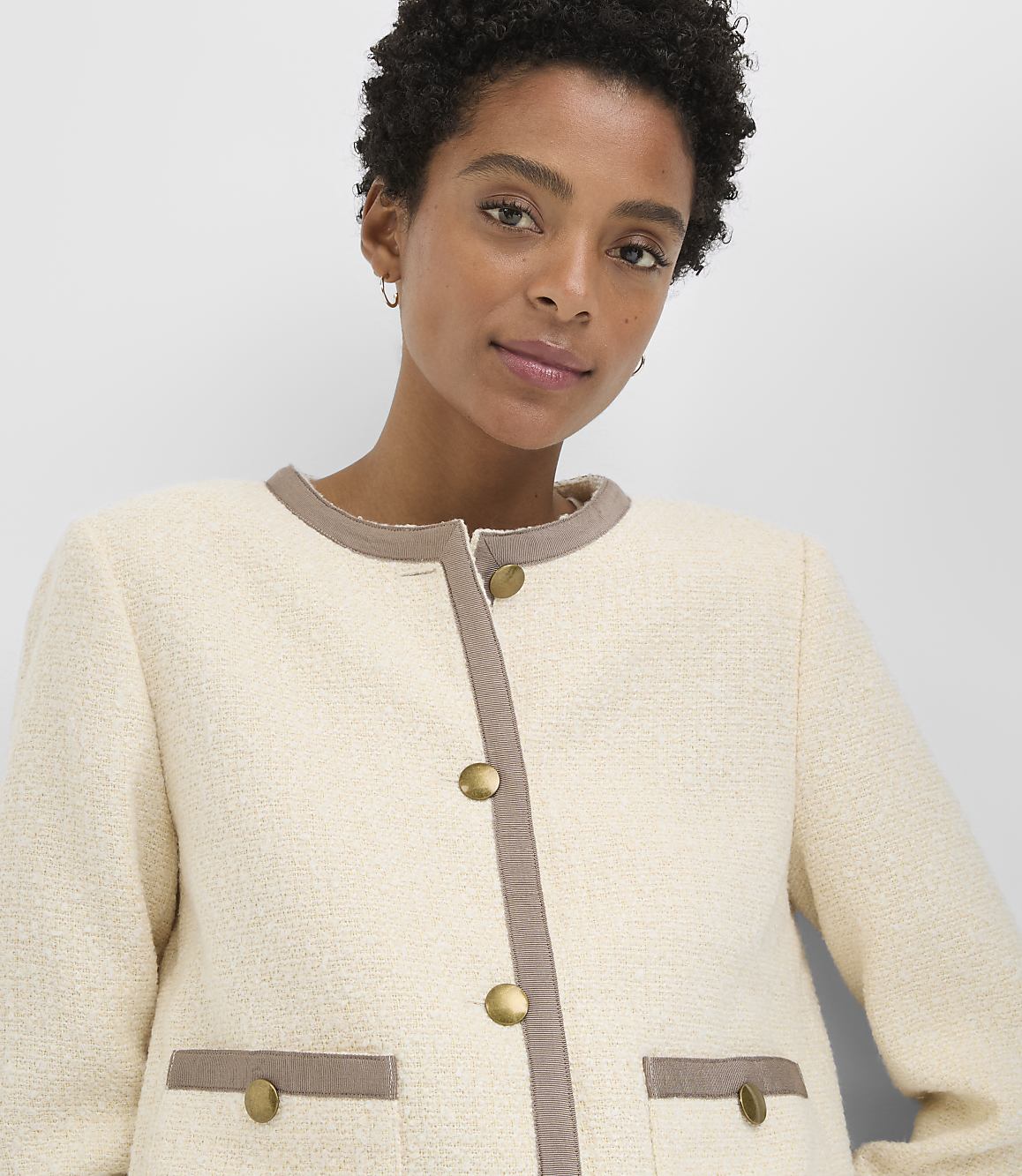 A woman in a cream-colored tweed jacket with brown trim and gold buttons stands against a plain gray background.