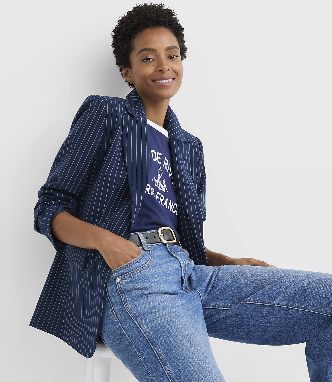 A woman in a pinstripe blazer and jeans smiles against a plain white background.