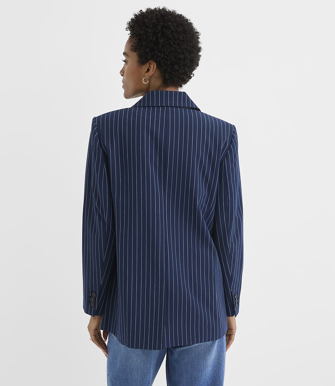 A woman in a navy pinstripe blazer and jeans stands against a plain white background.