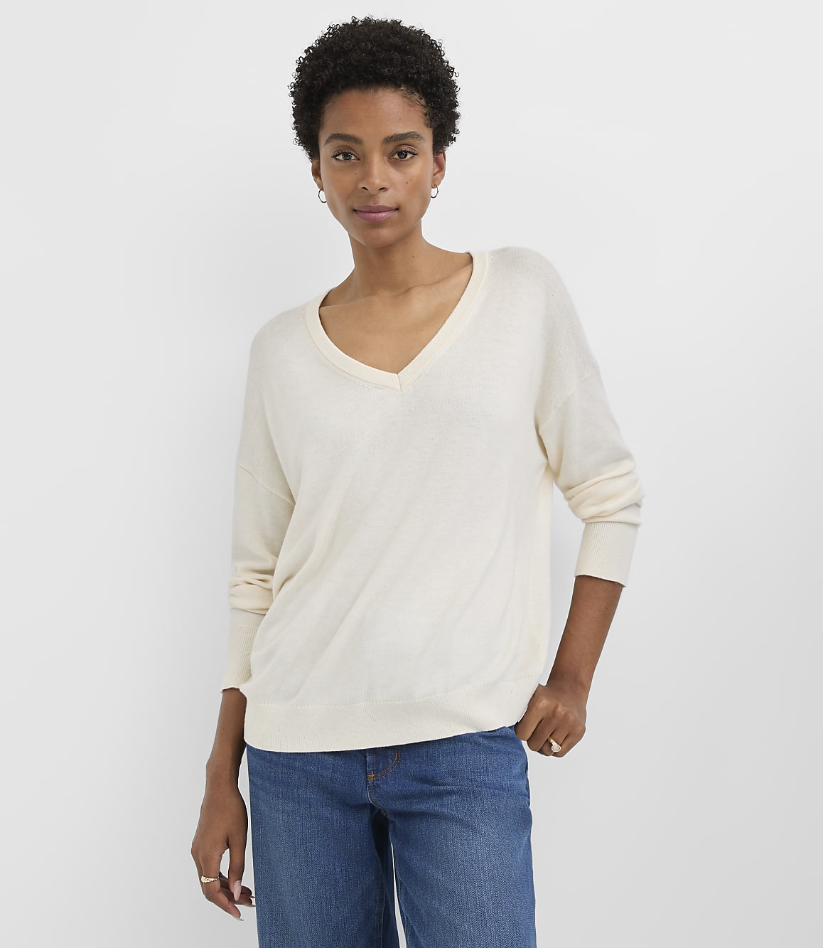 A woman in a cream v-neck sweater and blue jeans stands against a plain white background.