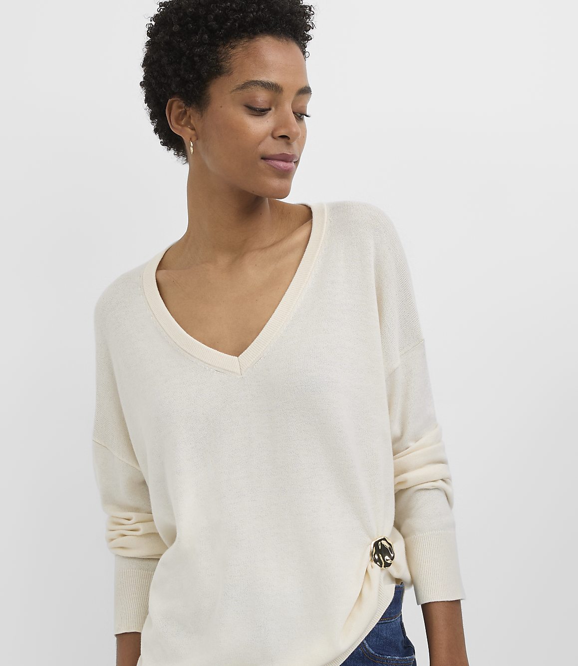 A woman in a cream v-neck sweater with a decorative button on the side stands against a plain white background.