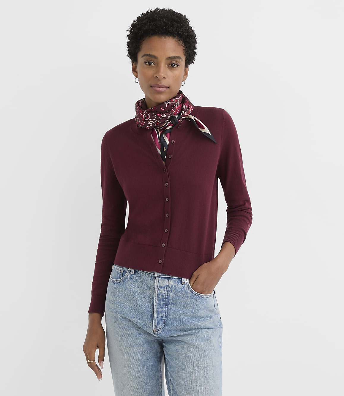 A woman in a burgundy cardigan and jeans stands against a plain white background.