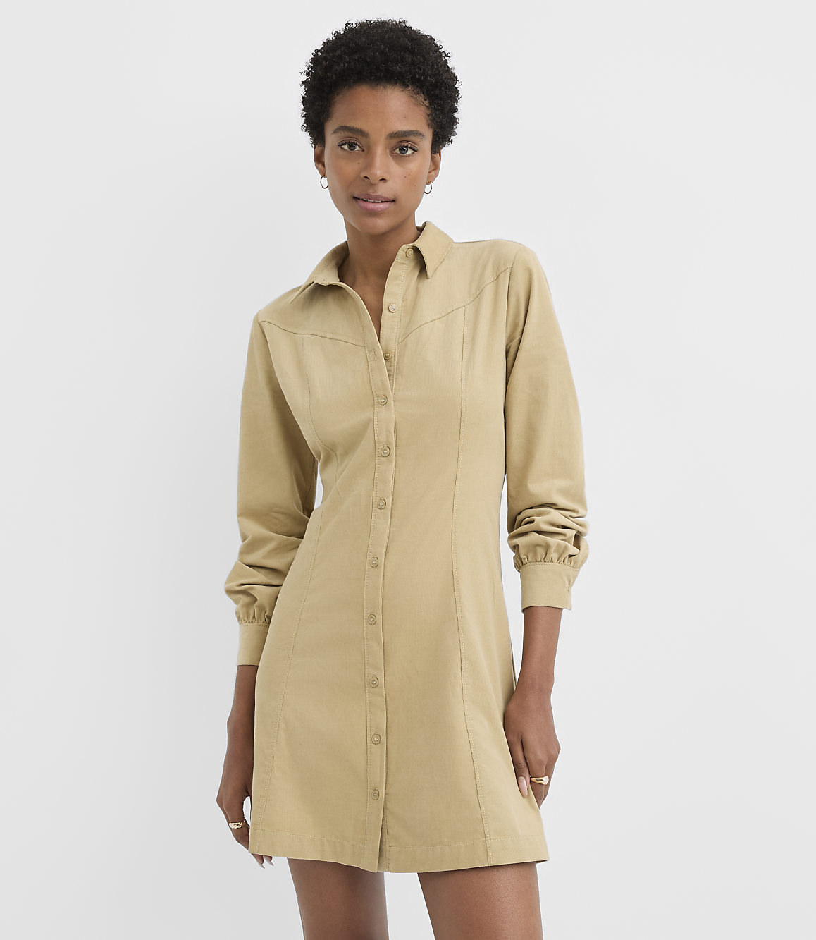 A woman in a tan button-down dress stands against a plain white background.