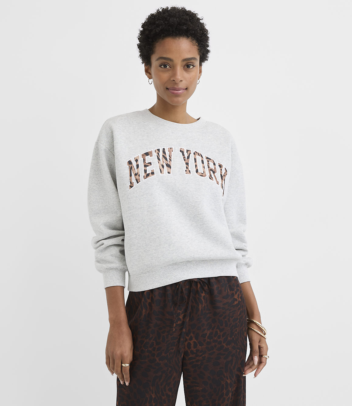 A woman in a gray sweatshirt with "NEW YORK" in leopard print stands against a plain gray background.