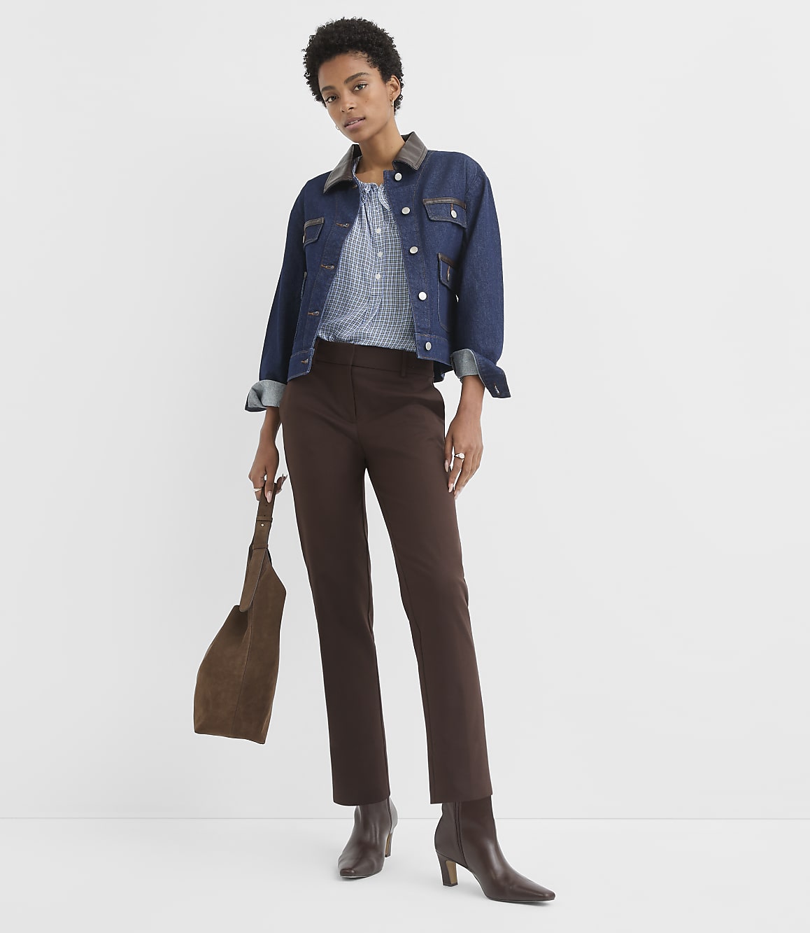 A woman in a denim jacket and brown pants stands against a plain white background, holding a brown suede purse.