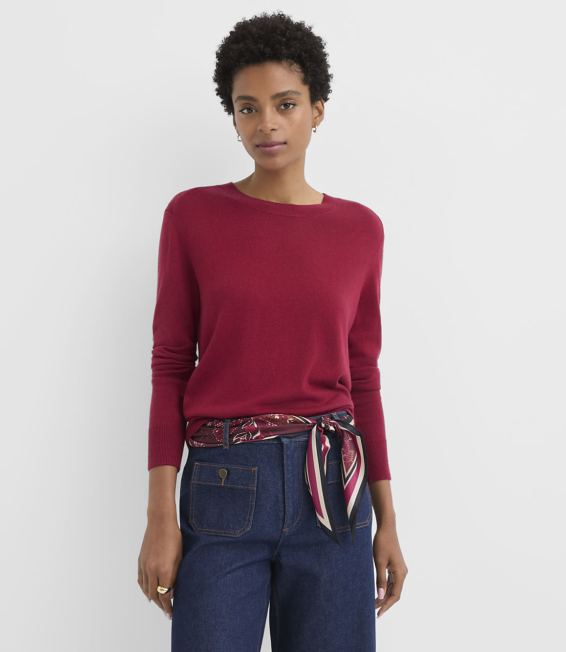 A woman in a red sweater and dark jeans stands against a plain white background.