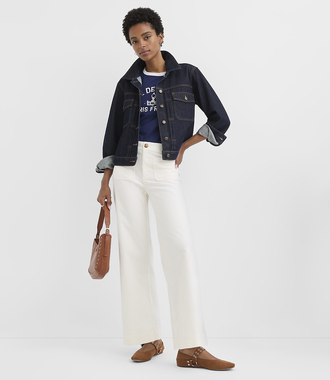 A woman in a denim jacket and white pants stands against a plain white background, holding a brown purse.