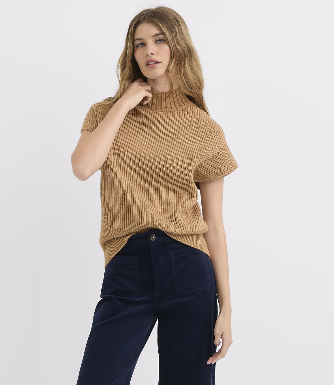 A woman in a tan mock-neck sweater and navy pants stands against a plain white background.