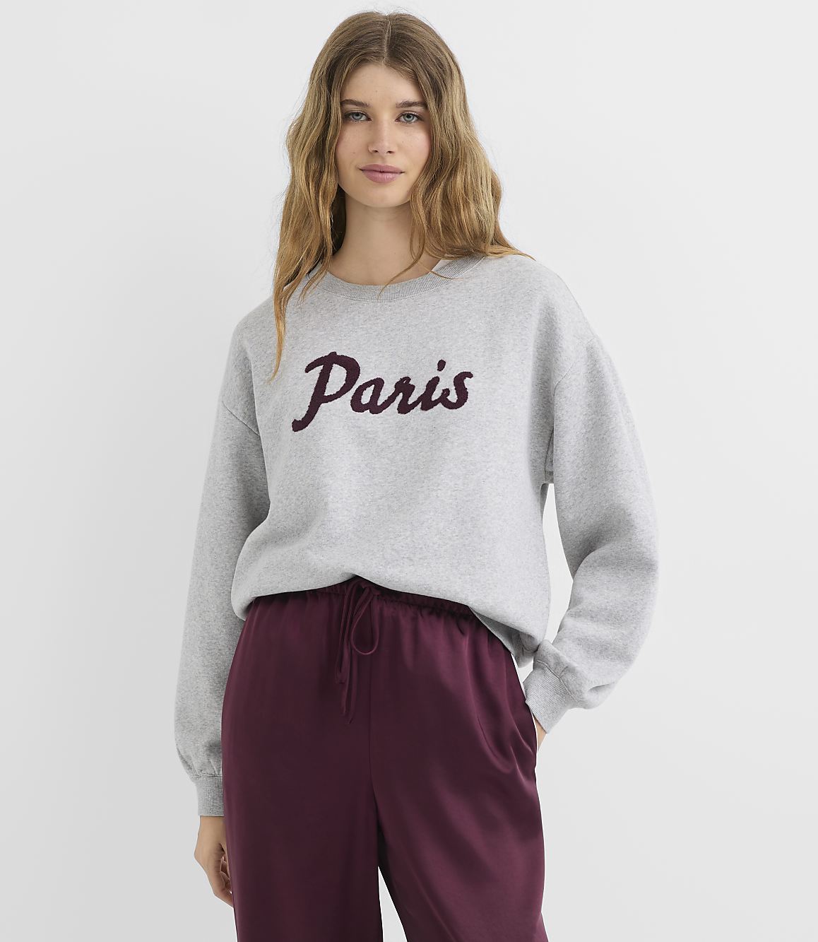 A young woman in a gray sweatshirt with "Paris" written on it stands against a plain white background.