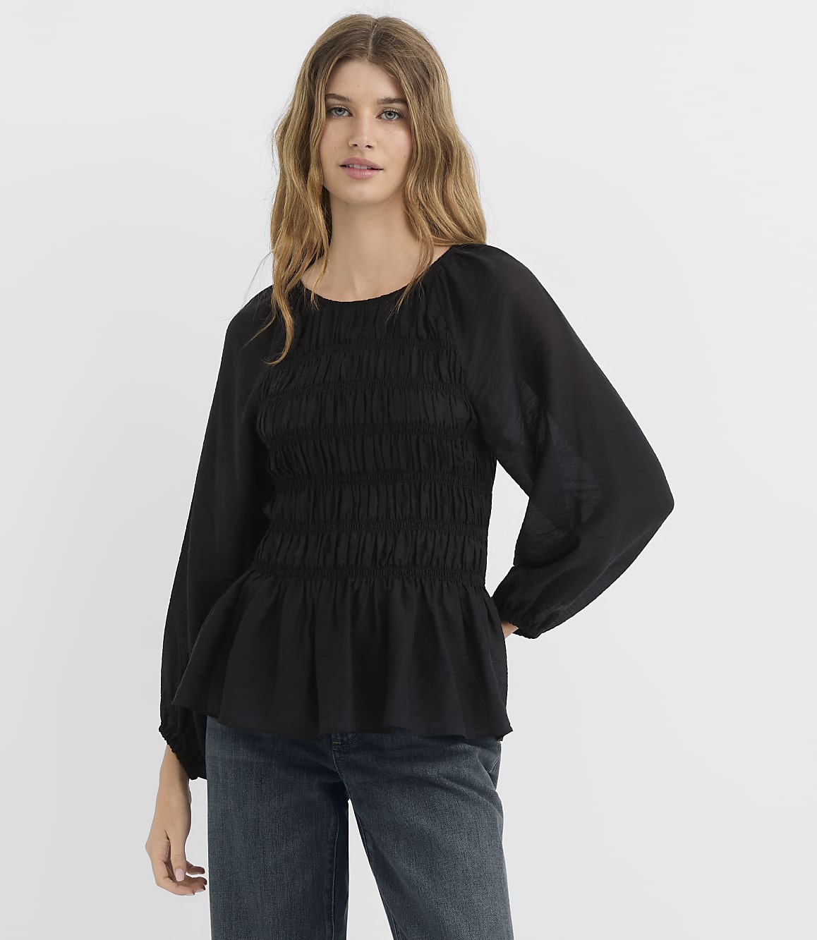 A woman in a black smocked-bodice blouse and jeans stands against a plain white background.