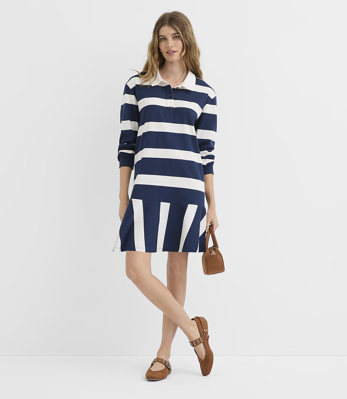 A woman in a navy and white striped polo dress stands against a plain white background, holding a small brown purse and wearing brown flats.