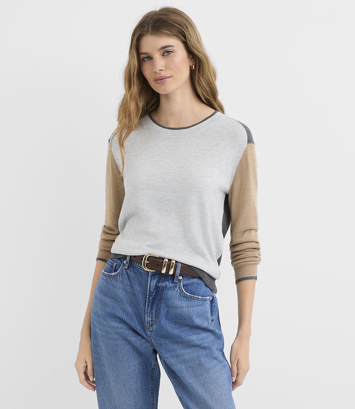 A woman in a color-blocked sweater and jeans stands against a plain gray background.